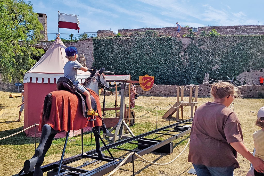 Ein Kind auf einem Spielgeräte-Pferd auf einem Spielplatz, im Hintergrund eine alte Schlossmauer.