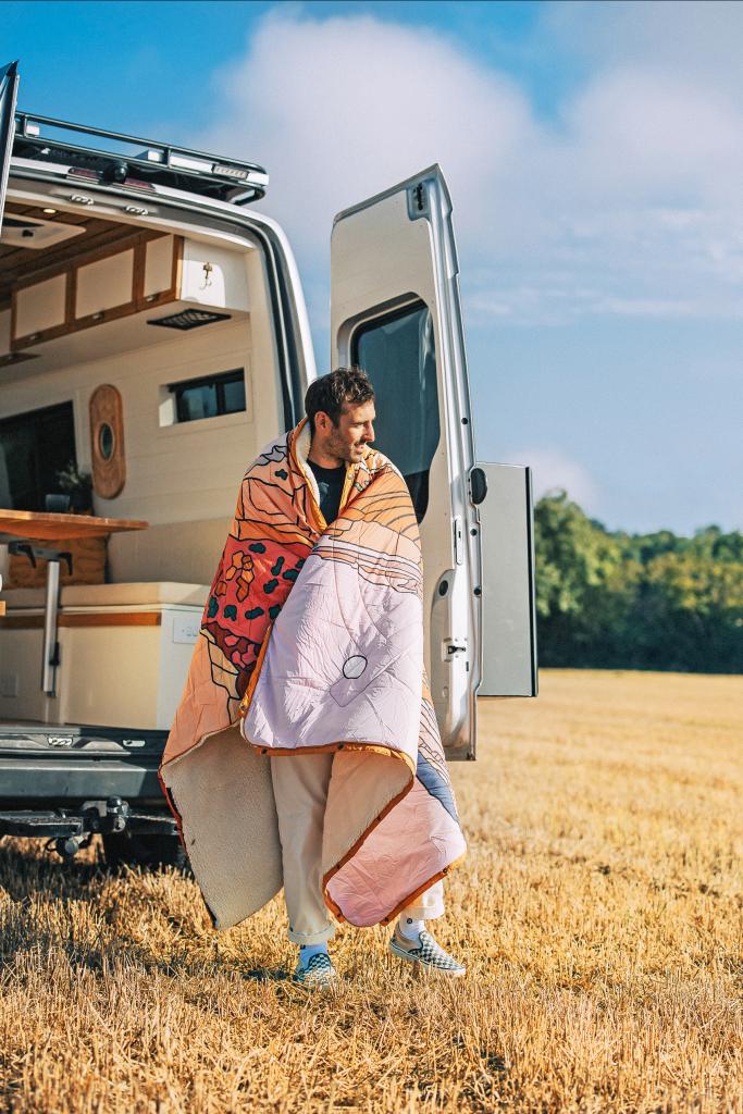 Mann vor einem Camper ist in eine multifunktionale Campingdecke von Voited eingehüllt