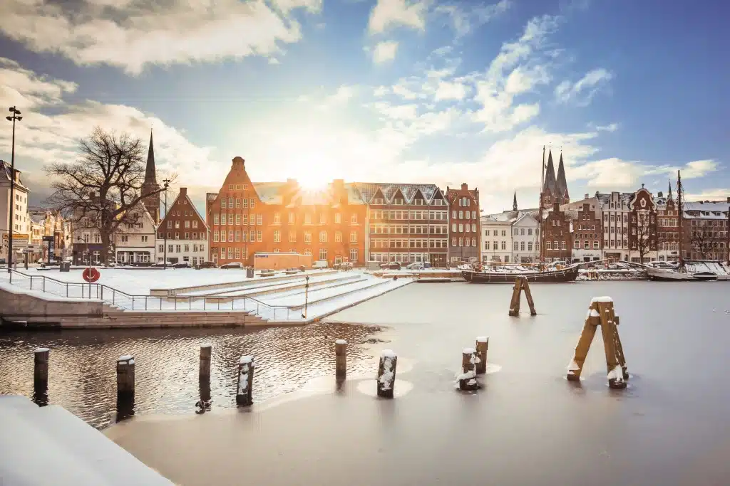 Winterliche Szene am Drehbrückenplatz in Lübeck, mit einer schneebedeckten Landschaft im Vordergrund. Die tiefstehende Sonne sendet ihre Strahlen über die traditionellen, giebelständigen Backsteingebäude, während im Hintergrund die spitz zulaufenden Türme der Stadt emporragen. Das gefrorene Wasser reflektiert das Sonnenlicht, und ein paar hölzerne Poller ragen aus dem Eis heraus, was der friedlichen, verschneiten Szene einen Hauch von Stille verleiht.