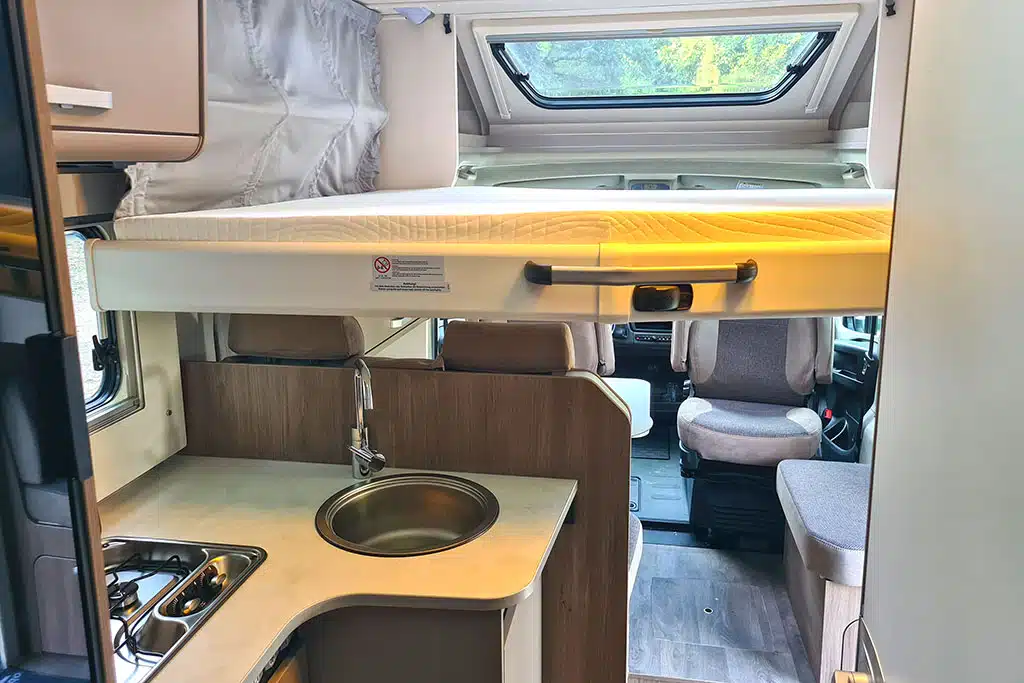 Carado T328 Pro+: Wohnraum im Wohnmobil mit herabgelassenem Hubbett, Sitzgruppe vorne und L-Küche dahinter