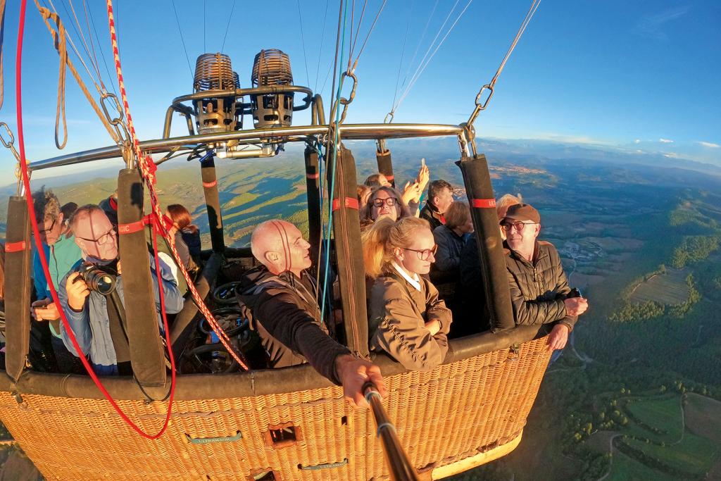 Menschen im Korb eines Heißluftballons, der über einer grünen, hügeligen Landschaft schwebt